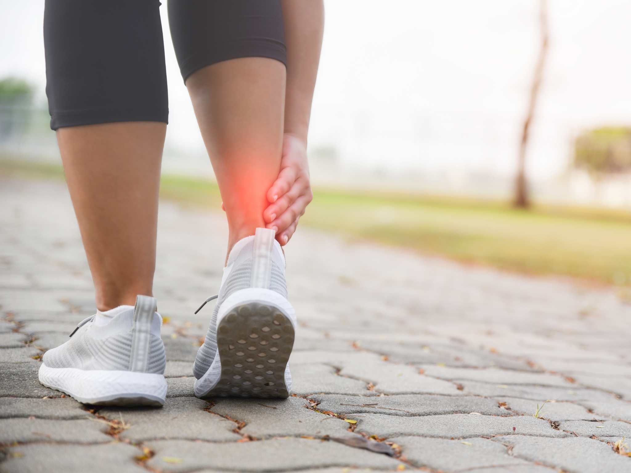 Woman hands holding her ankle while running on road in the park with red color highlight at ankle. Injury from workout concept.